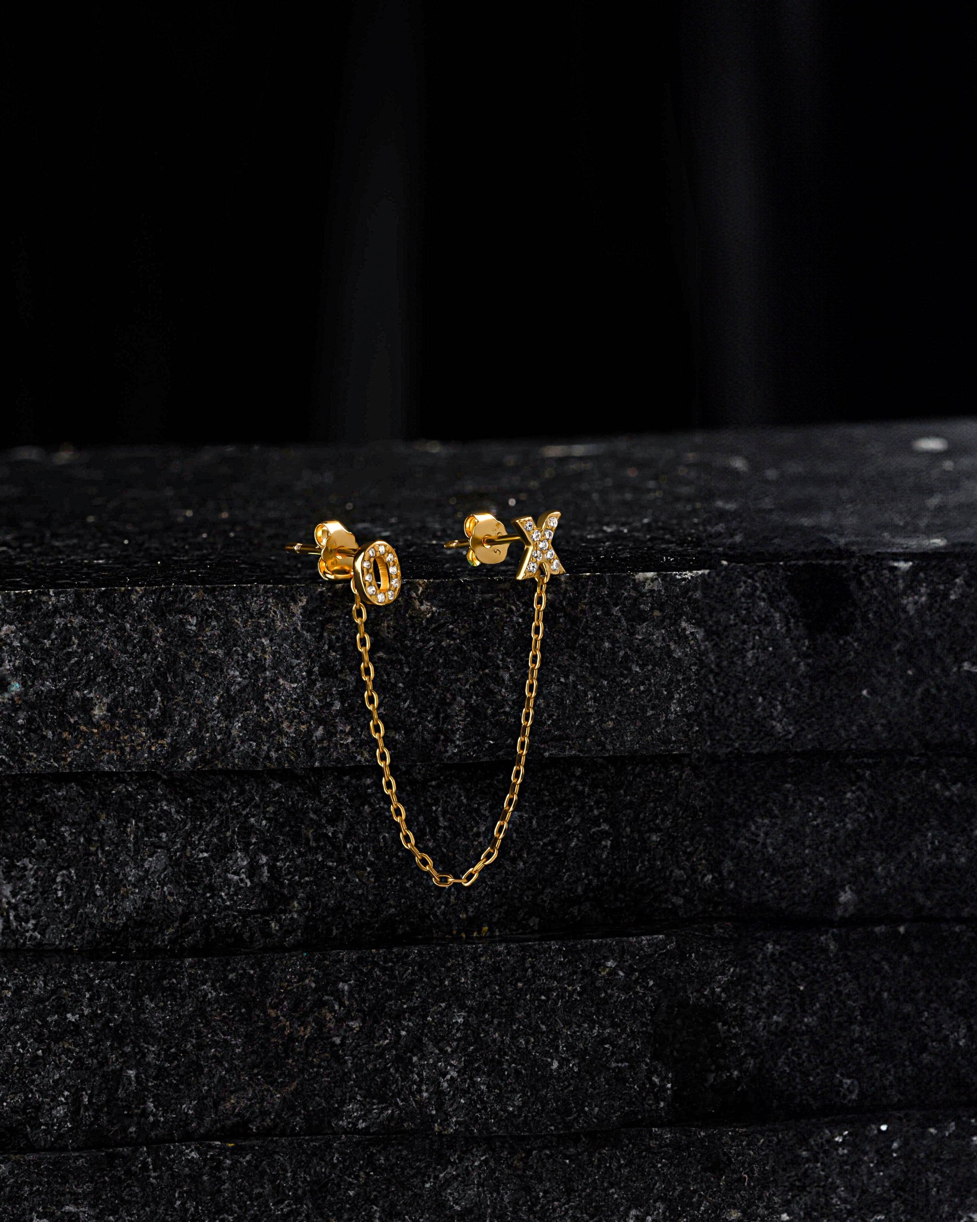 XO - Create 3 Chain Connector Earring displayed on black stone, showcasing gold chain and crystal-studded X and O studs for versatile styling.